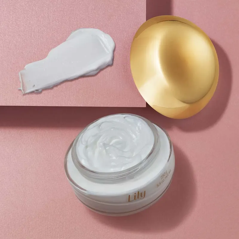 Creme Lily Essence aberto mostrando textura cremosa branca acetinada com tampa dourada ao lado sobre fundo rosa.